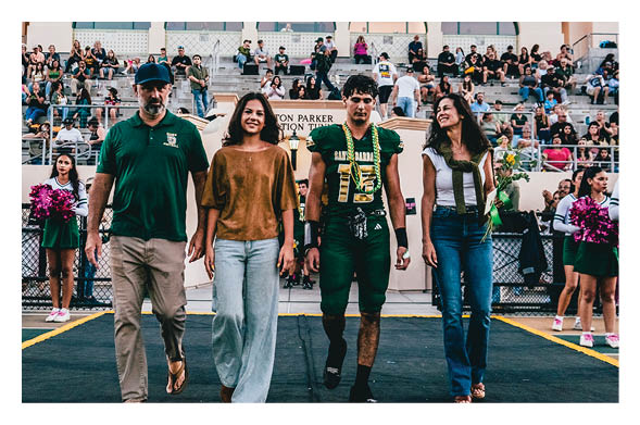 Four people walk side by side on a sports field with a crowd in the bleachers behind them and cheerleaders holding pom-poms on either side. Description generated by AI