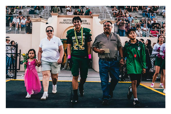 Five people, including a child in a pink dress and adults in casual and sports attire, walk hand in hand on a sports field with spectators seated in the background. Description generated by AI