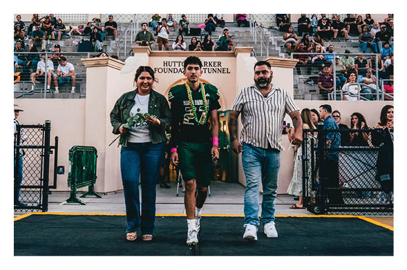 Three adults walk confidently toward the camera from a tunnel entrance with a crowd seated in bleachers behind them. Description generated by AI