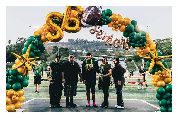 Five people stand together on a sports field under a decorative balloon arch with gold and green balloons, large gold letters “SB," a football balloon, and the word "Seniors" in cursive balloons. Description generated by AI