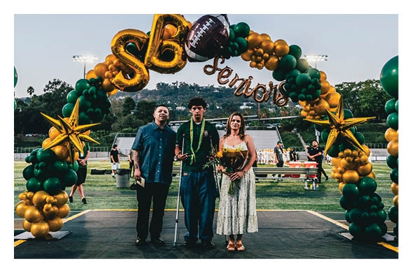 Three people stand together under a balloon arch decorated with gold and green balloons, large gold letters spelling “SB," a football balloon, and the word "Seniors," with one person using crutches and another holding flowers on a sports field. Description generated by AI