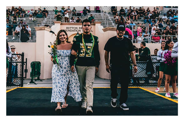 A person wearing a sports jersey walks arm-in-arm with another person holding flowers while a third person walks beside them on a field with spectators seated in the background. Description generated by AI