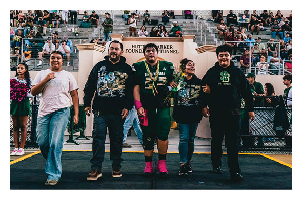 Five people stand side by side on a sports field with a crowd seated in bleachers behind them, and two people on each side holding pink pom-poms. Description generated by AI