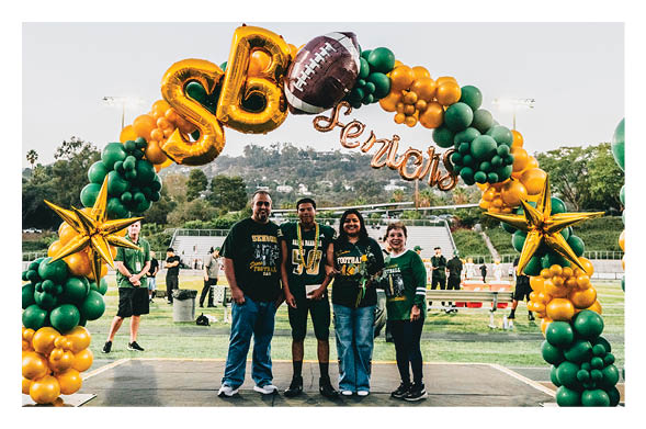Four people standing together smiling under a green and gold balloon arch decorated with stars and a football balloon with the word “Seniors" at a sports field. Description generated by AI