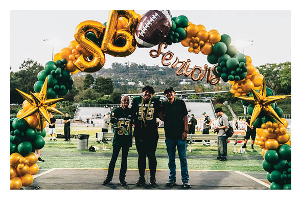 Three people stand smiling under a colorful balloon arch decorated with gold stars, green and gold balloons, and letters spelling “SB Senior" on a sports field with bleachers and trees in the background. Description generated by AI