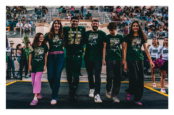 People wearing matching sports-themed shirts walk together on a track with a crowd seated in bleachers behind them. Description generated by AI