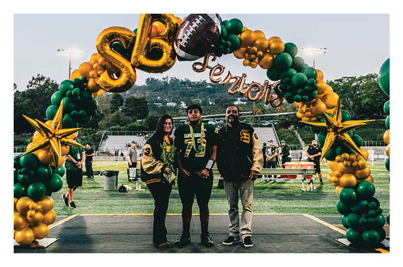 Three people wearing sports jackets stand under a decorative balloon arch with gold and green balloons, gold stars, and a football balloon on a sports field. Description generated by AI