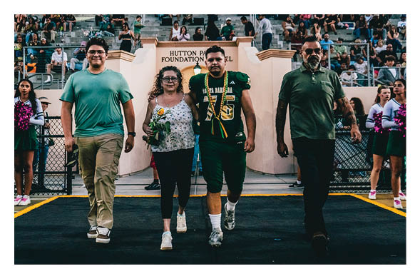 Four people walk arm in arm on a football field with a crowd in the stands behind them and cheerleaders on either side. Description generated by AI