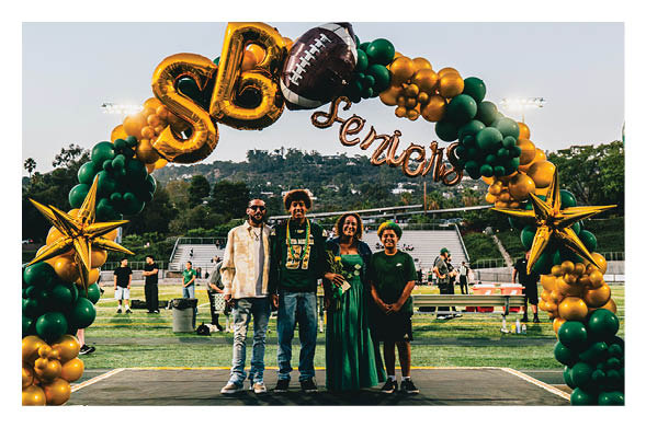 Four people stand smiling under a green and gold balloon arch decorated with a football and the word seniors on a sports field. Description generated by AI