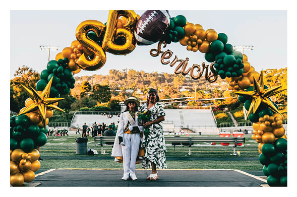 Two people stand on a stage under a green and gold balloon arch decorated with football and star-shaped balloons, with a sports field and bleachers in the background. Description generated by AI