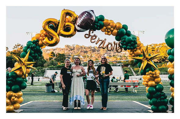 Four people stand smiling under a decorative balloon arch with green and gold balloons, large gold stars, and football-themed decorations on a sports field during a senior celebration event. Description generated by AI