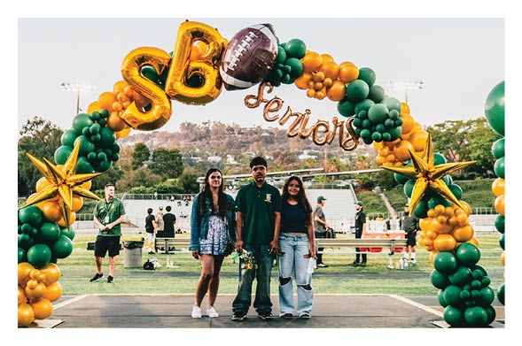 Three people stand together smiling under a green and gold balloon arch decorated with a football and the words “SB Seniors" at an outdoor event on a sports field. Description generated by AI