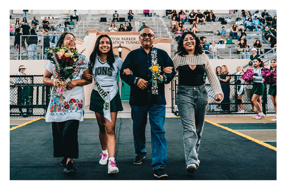 Four people smiling and walking arm in arm on a sports field with one person wearing a cheerleading uniform and others holding flowers while spectators watch from bleachers in the background. Description generated by AI