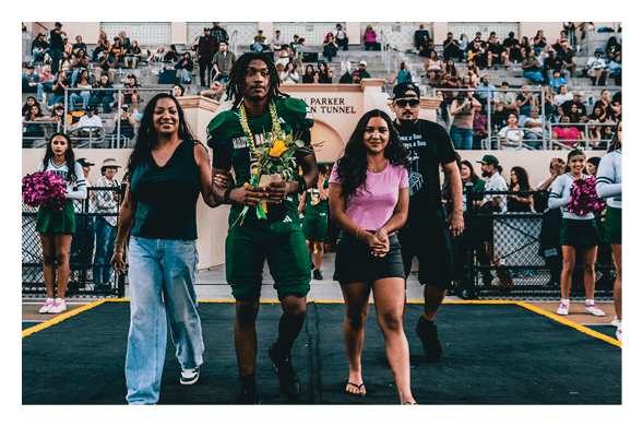 Four people walk together on a sports field with a crowd seated in bleachers behind them, one person holding flowers and wearing a green sports uniform. Description generated by AI