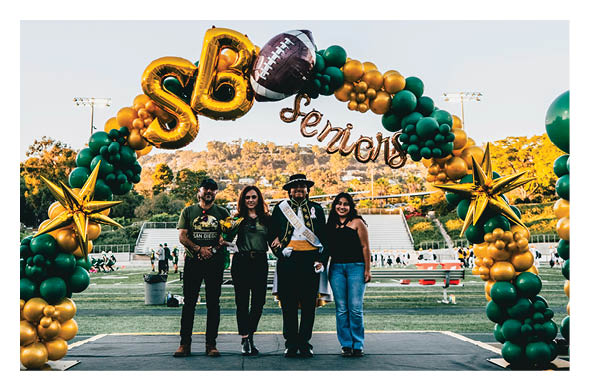 Four adults stand smiling under a green and gold balloon arch decorated with a football and the letters SB and the word seniors on a sports field. Description generated by AI