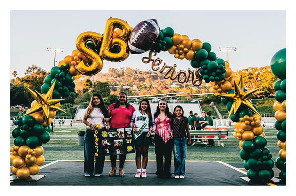 Five people stand smiling under a green and gold balloon arch decorated with large star balloons and football-themed balloons on a sports field. Description generated by AI