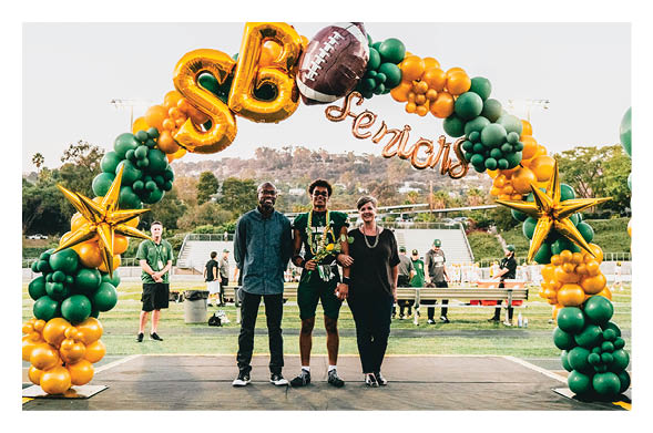 Three people stand together on a stage under a green and yellow balloon arch decorated with gold stars and a football balloon, with a sports field and spectators in the background. Description generated by AI