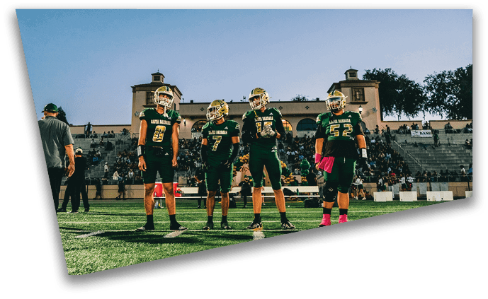 Four people in football uniforms stand on a field with a stadium and spectators in the background during a clear day. Description generated by AI