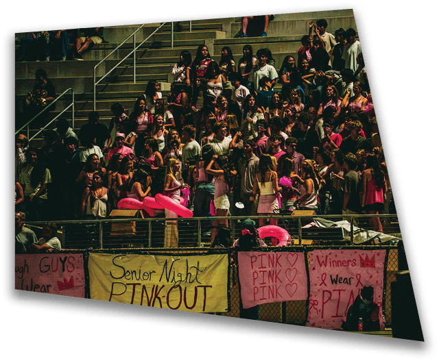 Banners with messages supporting wearing pink for Senior Night Pink Out are displayed on a railing at a nighttime event with people standing behind them. Description generated by AI
