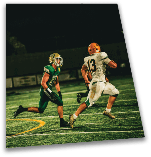 Two people in football uniforms are running on a field at night, with one person in white and orange running ahead and the other in green and gold chasing behind. Description generated by AI