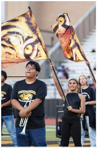A person wearing a black sleeveless top and black pants is smiling and holding up a flag with a tiger pattern while standing on a sports field with other people in the background. Description generated by AI