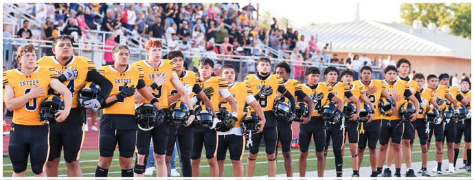 A line of people in yellow and black football uniforms stand side by side on a field with a crowd seated in bleachers behind them. Description generated by AI