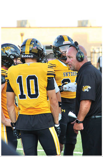 A coach wearing a headset is talking to two football players in yellow and black uniforms on a field. Description generated by AI