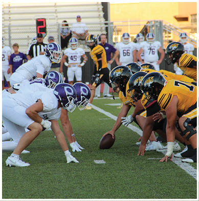 Two football teams in white and yellow uniforms are lined up facing each other on a green field preparing for a play with players in helmets and pads. Description generated by AI