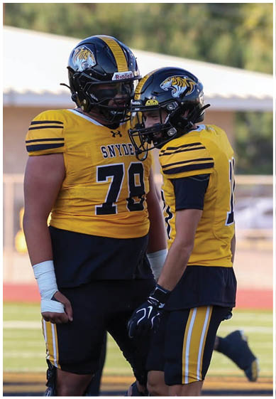 Two people wearing yellow and black football uniforms with helmets stand on a field, one with a wrapped wrist. Description generated by AI