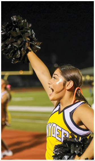 A person in a yellow and black cheerleading uniform is enthusiastically raising a black pom-pom with glitter on their face at a nighttime sports event. Description generated by AI