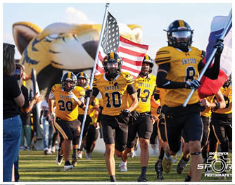 People in yellow and black football uniforms run onto a field holding American and Texas flags with a large inflatable tiger head in the background. Description generated by AI