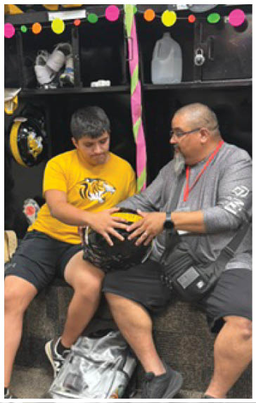 Two people sitting closely on a bench indoors, both holding a black and yellow helmet, with one wearing a yellow shirt and the other wearing a gray shirt and a crossbody bag. Description generated by AI