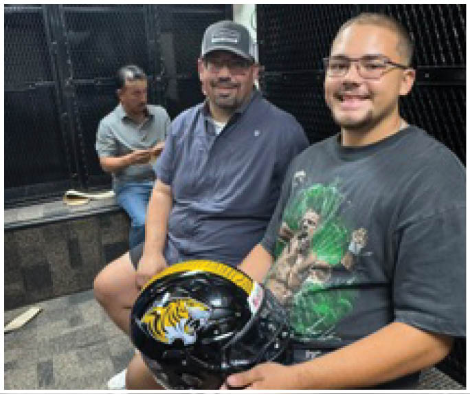 Three adults sit indoors on a bench, with one holding a black helmet featuring a yellow and white tiger design. Description generated by AI