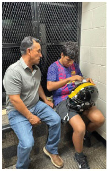 Two people sit on a bench in a room with cinder block walls, one wearing a gray polo and jeans and the other wearing a colorful sports jersey and shorts while holding a black helmet with yellow tape. Description generated by AI