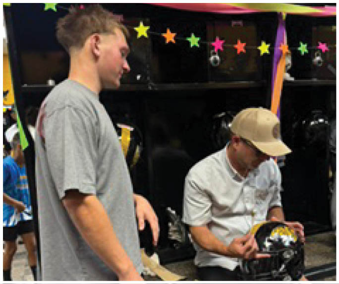 Two adults are in a locker room with one sitting on a bench holding a football helmet while the other stands nearby. Description generated by AI