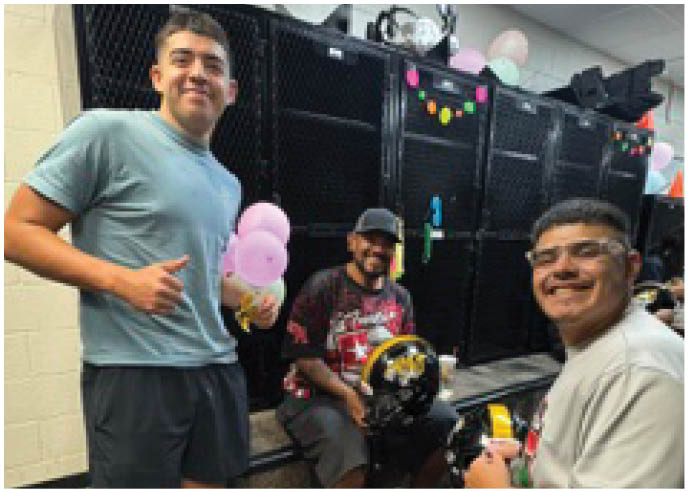 Three adults are smiling and posing together indoors with one holding pink balloons and another holding a yellow object. Description generated by AI