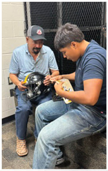 Two people are sitting indoors near a black metal locker, with one holding a black football helmet while the other appears to be signing or writing on it. Description generated by AI