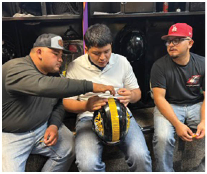 Three adults sit closely together on a bench with one person holding a black and yellow helmet while another points at it. Description generated by AI