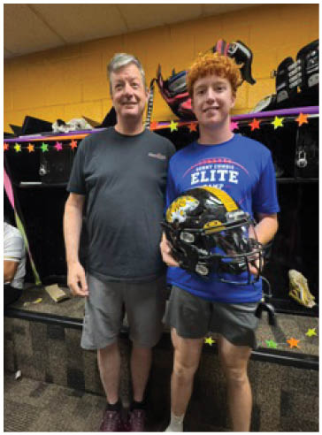 Two people stand side by side indoors with one holding a football helmet and both smiling, surrounded by colorful star decorations. Description generated by AI