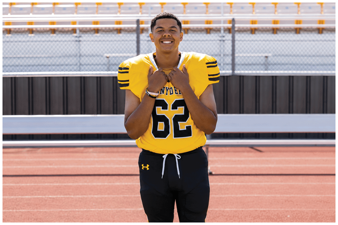 A person wearing a yellow football jersey with the number 62 and black shorts is standing on a sports field with a running track and bleachers in the background. Description generated by AI