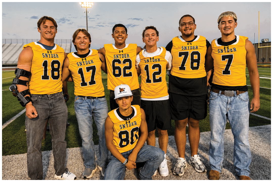 Six adults wearing yellow Snyder football jerseys and casual pants pose together on a football field with one adult kneeling in front wearing a white cap. Description generated by AI
