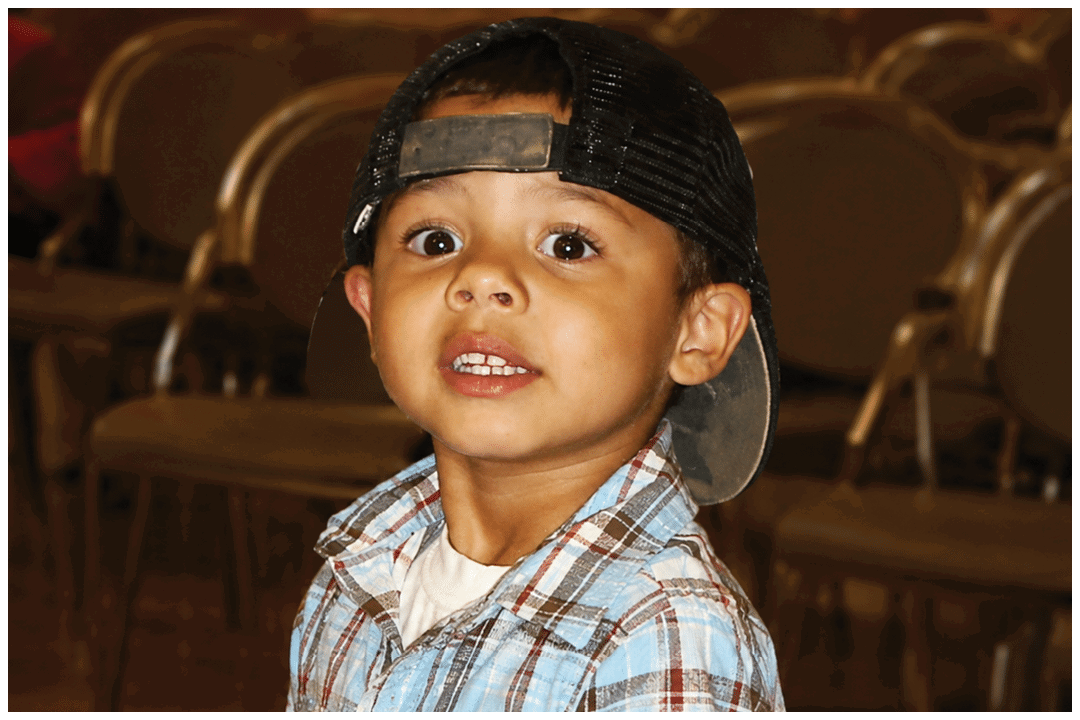 A child wearing a backward baseball cap and a blue plaid shirt looks slightly upward with a neutral expression in a room with wooden floors and chairs. Description generated by AI