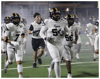 People in yellow and black football uniforms are on a field with one player in a white and purple uniform facing them. Description generated by AI