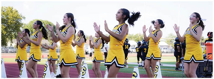 A line of people in yellow and black football uniforms stand side by side on a field with a crowd seated in bleachers behind them. Description generated by AI