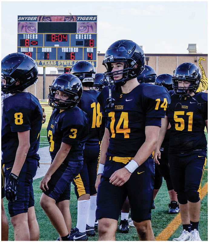 Two people in yellow and black football uniforms are playing against opponents in white and purple uniforms on a football field with spectators in the background. Description generated by AI