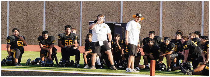A line of people in yellow and black football uniforms stand side by side on a field with a crowd seated in bleachers behind them. Description generated by AI
