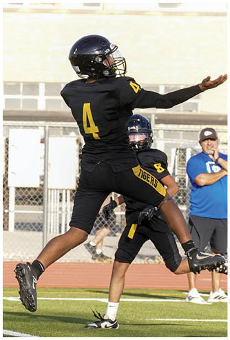 A coach wearing a headset is talking to two football players in yellow and black uniforms on a field. Description generated by AI