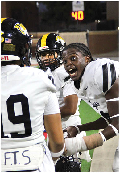 Three people in football uniforms and helmets are engaged in an animated conversation on a field with a scoreboard in the background. Description generated by AI