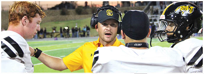A person wearing a yellow shirt and headset is talking to three people in football uniforms on a field. Description generated by AI