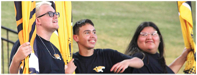 Three people wearing black shirts with a tiger logo are holding yellow and black flags and smiling outdoors. Description generated by AI